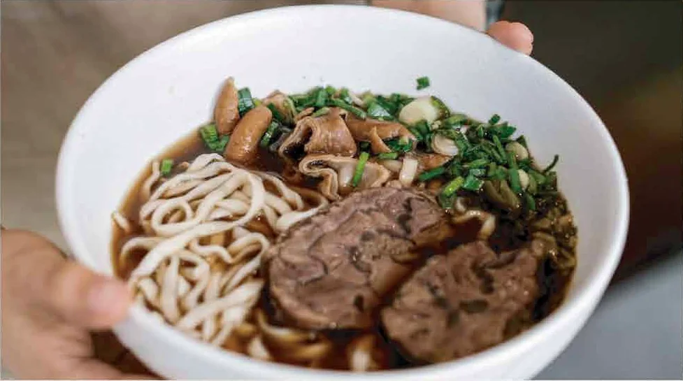 A hand holding a white bowl of Grandma Lin's beef noodle soup with wide noodles, beef shank, pickled greens and scallions